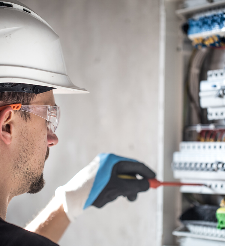 Ein Elektriker mit Schutzhelm, Schutzbrille und Handschuhen arbeitet an einem Sicherungskasten.