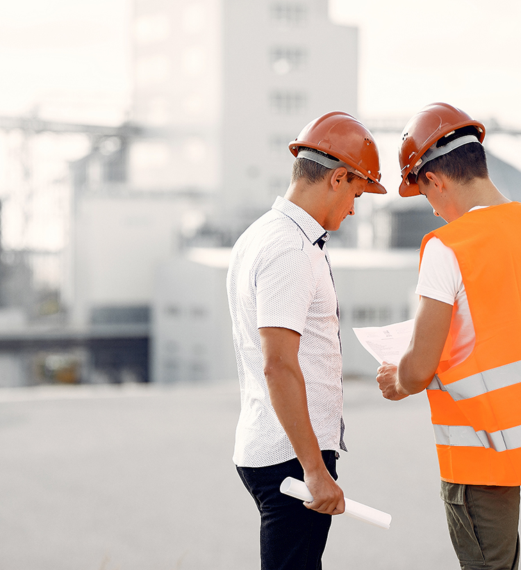 Zwei Männer mit orangen Schutzhelmen und einem Bauplan im Freien, im Hintergrund unscharfe Industriegebäude