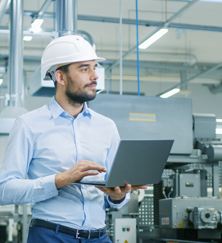 Ein Ingenieur in einem hellblauen Hemd und weißem Schutzhelm hält einen Laptop in einer technischen Anlage.