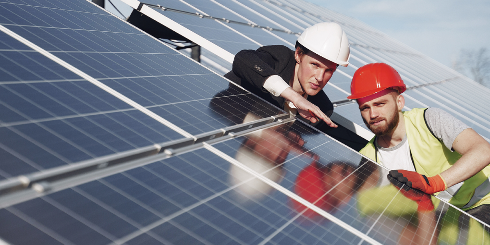 Zwei Männer mit Schutzhelmen überprüfen Solarpanels auf einem Dach, einer trägt weißen Helm und Anzug, der andere roten Helm, gelbe Sicherheitsweste und Handschuhe.