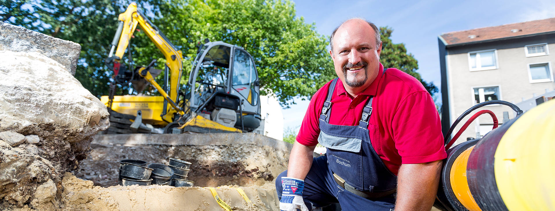 Ein Arbeiter der Stadtwerke Bochum Netz kniet an einer Baustelle im Freien neben einem großen gelben Rohr und einer Bagger-Maschine.