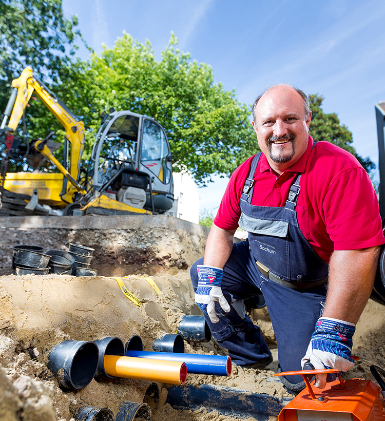 Ein Arbeiter der Stadtwerke Bochum Netz kniet auf einer Baustelle vor mehreren Rohrleitungen aus Kunststoff. Im Hintergrund steht ein kleiner Bagger vor einem Baum unter blauem Himmel.