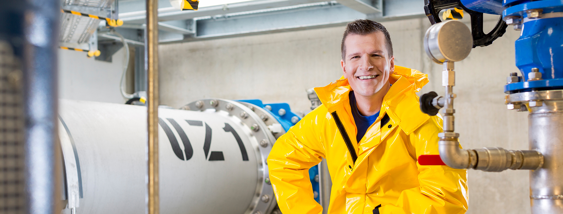 Ein lachender Mann in einem gelben Schutzanzug steht in einem technischen Raum mit großen Rohren und Ventilen.
