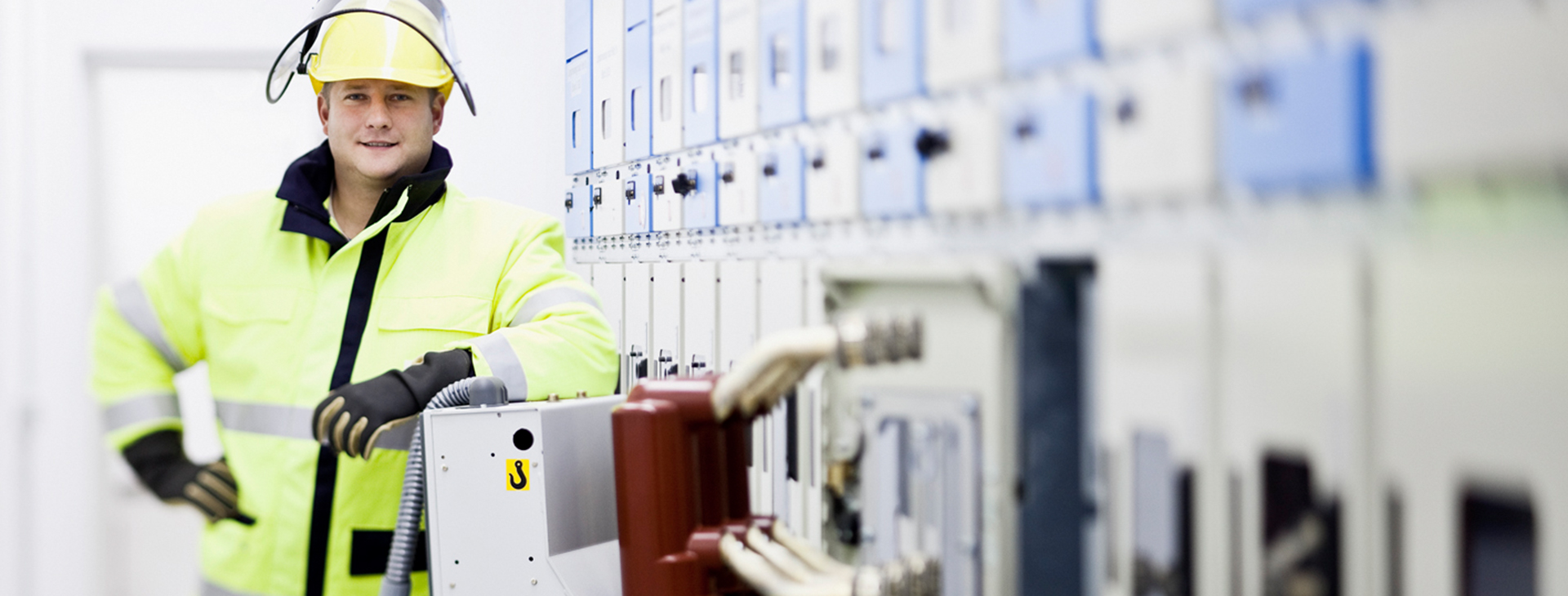 Ein Techniker in Schutzkleidung und Helm steht vor einer Wand mit elektrischen Schaltanlagen.