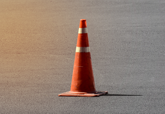 Ein oranger Baustellen-Absperrkonus mit zwei weißen Streifen steht auf frisch asphaltiertem Straßenbelag.